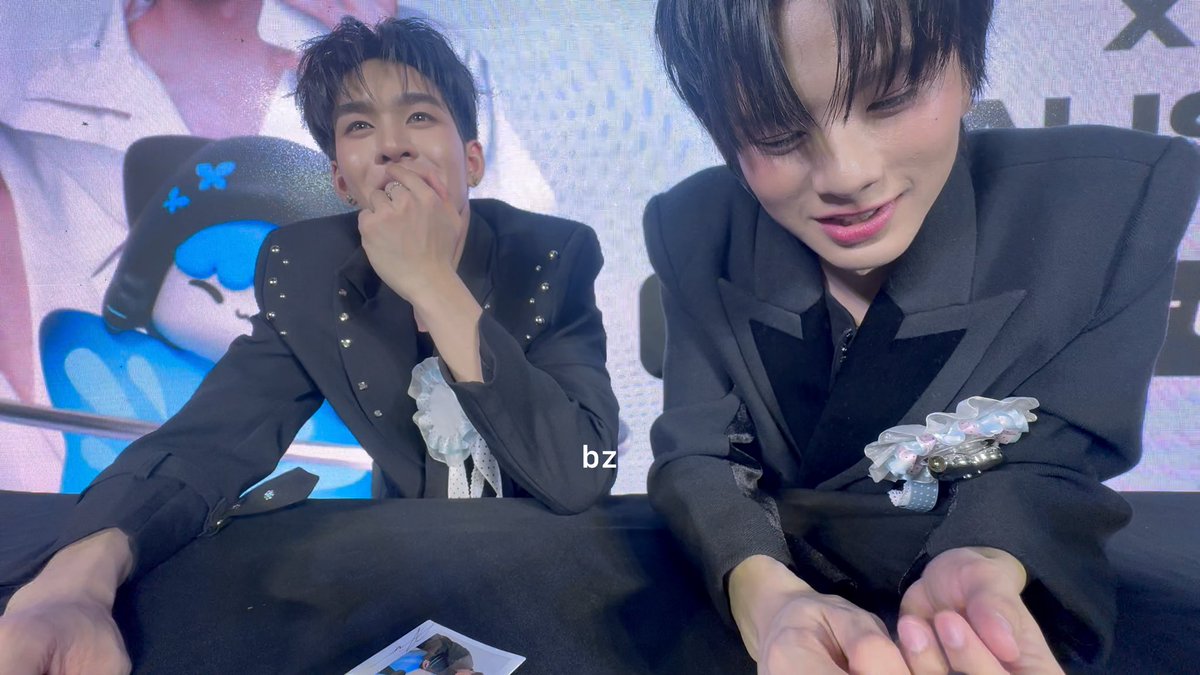 Two young men in matching black suits with embellished lapels pose at what appears to be a fan event, with a plush toy visible in the background and "bz" watermark.
