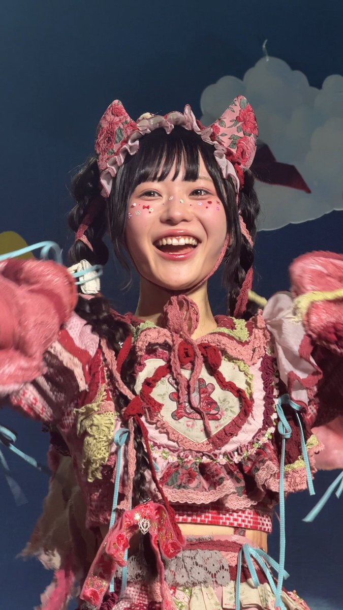 A smiling performer in an elaborate pink and red frilly costume with cat-ear-like head accessories and face stickers, waving pink pom-poms on stage.