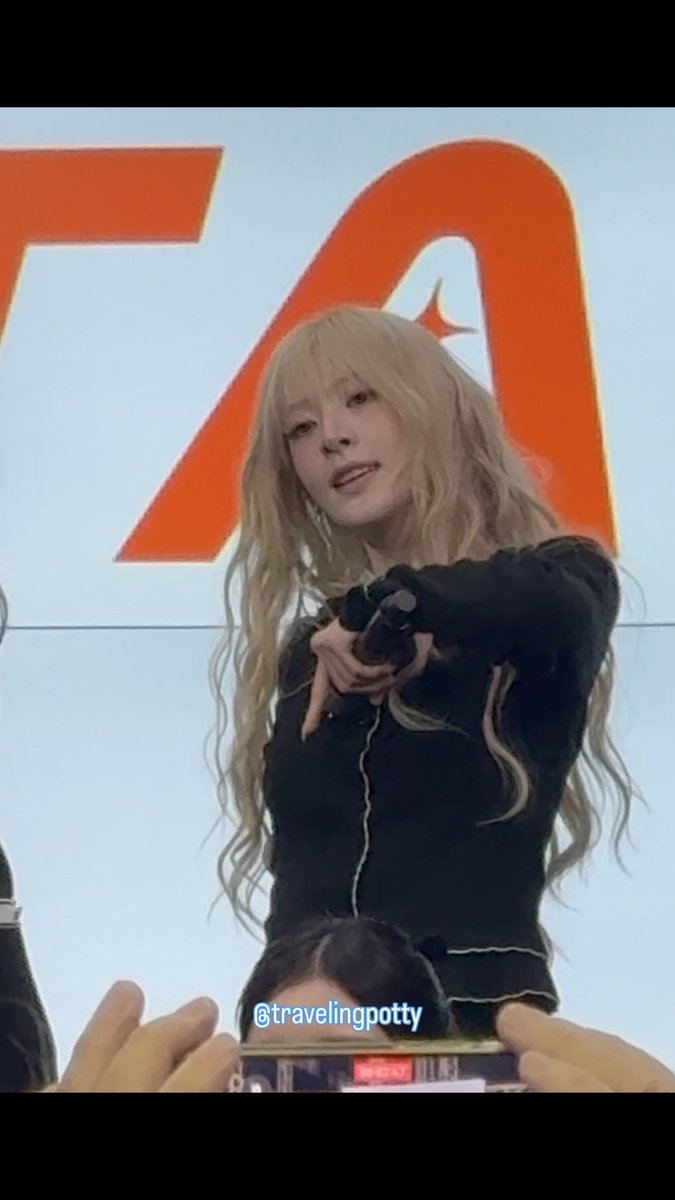 A blonde-haired woman in a black top holds a microphone at what appears to be a fan event, with a large orange logo visible in the background and the watermark '@travelingpotty'.