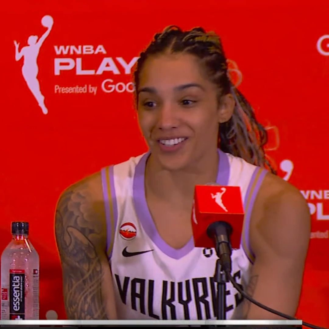 A female basketball player with braided hair, wearing a white and purple 'VALKYRIES' jersey, speaks at a press conference with a 'WNBA PLAYOFFS' backdrop.
