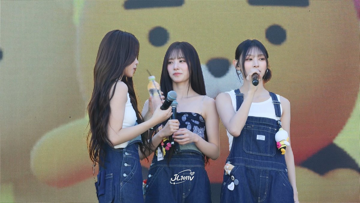 Three young female performers stand on stage in denim overalls and white tank tops, holding microphones, with the watermark 'JL10MV' visible.