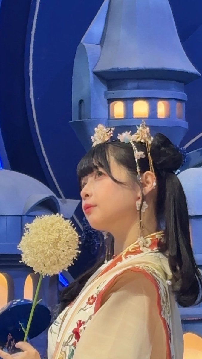 A young woman wearing ornate floral headwear and a flowing traditional-style dress holds a yellow pom-pom flower, posed against a fantasy castle backdrop.