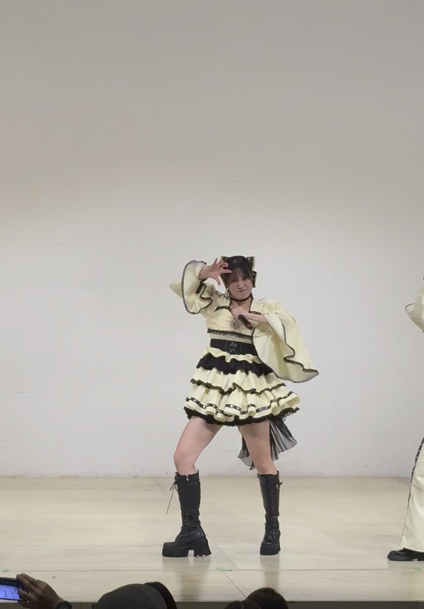 A young female performer on a stage poses with her hands near her face, wearing a yellow and black ruffled dress with a black belt, black knee-high platform boots, and cat-ear hair accessories.