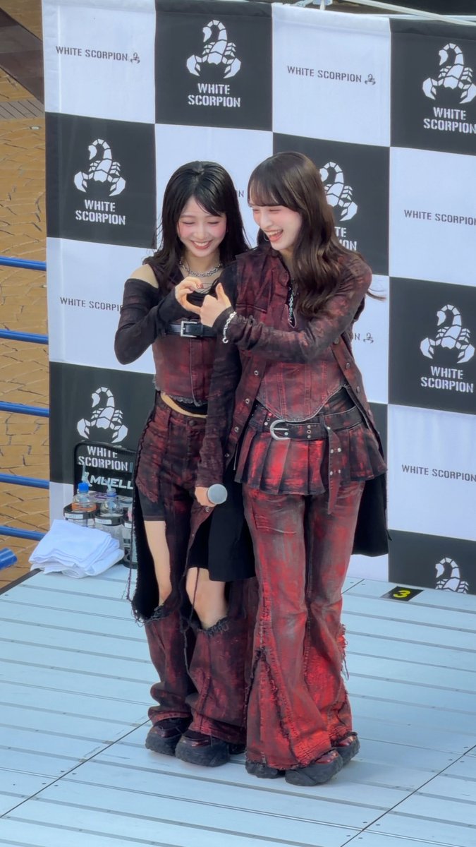 Two young women pose together making a heart shape with their hands at a 'WHITE SCORPION' event, wearing matching red and black denim outfits.