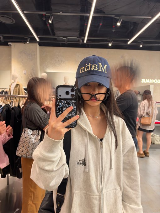 A mirror selfie of a woman wearing a navy 'MSLIA' cap, black glasses, and a white zip-up hoodie, taken with a phone featuring a black and white speckled case in a clothing store.