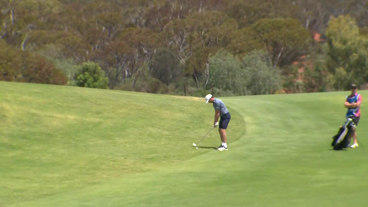A golfer in a blue shirt and dark shorts putts on a green fairway while another person with a golf bag stands to the side.
