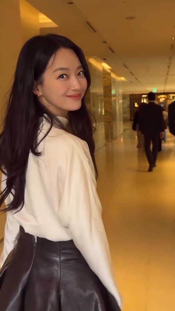 A young woman with long black hair, wearing a white blouse and black leather skirt, smiling as she looks over her shoulder in what appears to be a hotel hallway.
