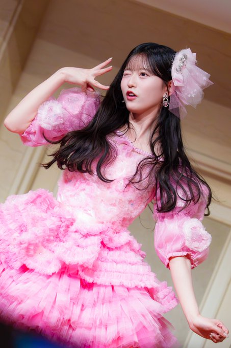 A young woman with long black hair wearing an elaborate pink ruffled dress and a large pink hair accessory, posing playfully.