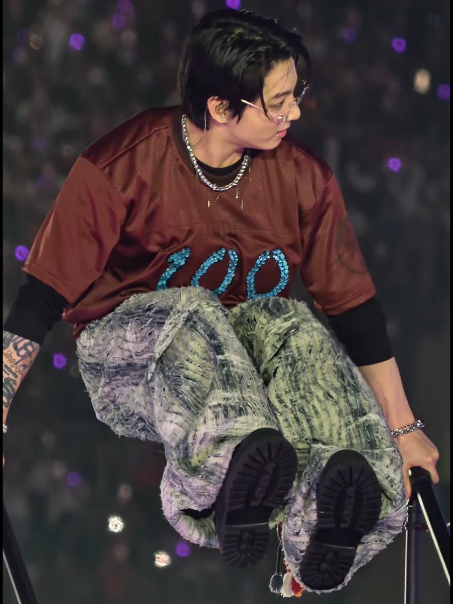 A performer wearing glasses, a brown shirt, and paint-splattered green pants sits on the edge of a stage during a concert, with their black UGG boots visible.