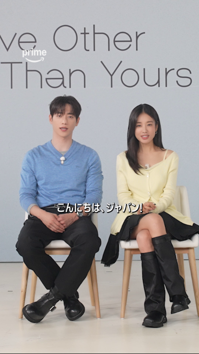 Two lead actors, Seo Kang-joon in a blue sweater and Ahn Eun-jin in a yellow cardigan, sit smiling on white chairs in front of a backdrop reading "Love Other Than Yours" with the Prime logo, greeting viewers with a Japanese subtitle "こんにちは、ジャパン！" ("Hello, Japan!"), promoting their new Korean romance series now streaming on Prime.