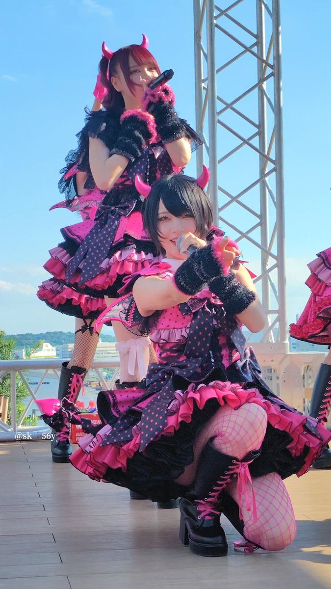 Two performers in elaborate pink and black gothic Lolita-style costumes with horn headpieces, posing on an outdoor stage.