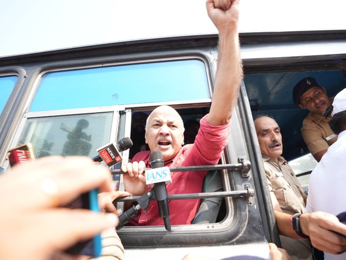 A man in a red shirt leans out of a vehicle window with his fist raised, speaking into microphones labeled 'IANS' and 'Zee Business' while surrounded by police and reporters.