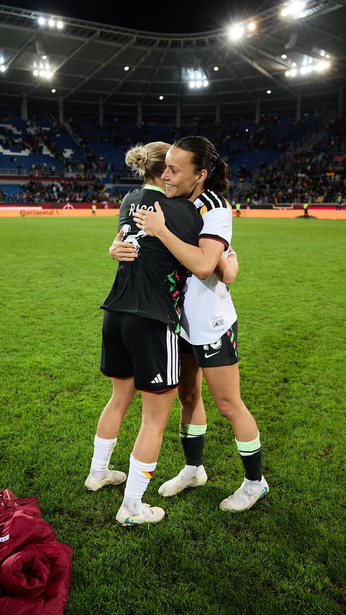 Two female soccer players in black and white uniforms hug on a green field inside a large stadium, appearing to celebrate.