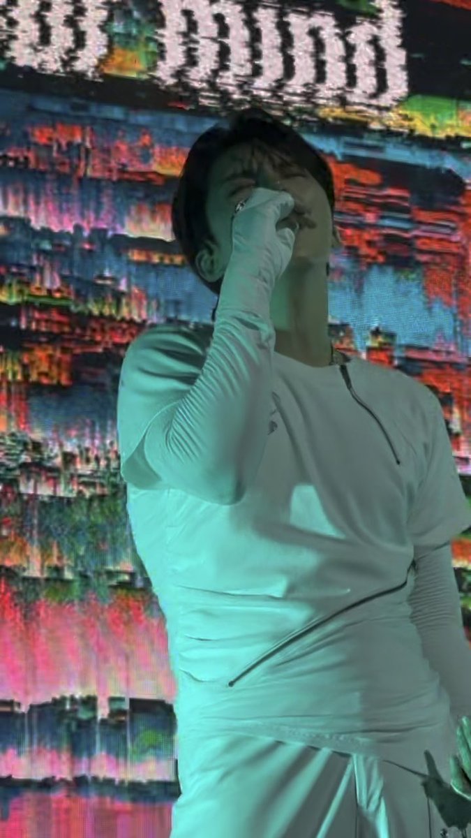 A male performer in a white long-sleeve outfit sings into a microphone on stage, with a colorful glitch-style digital screen glowing behind him.