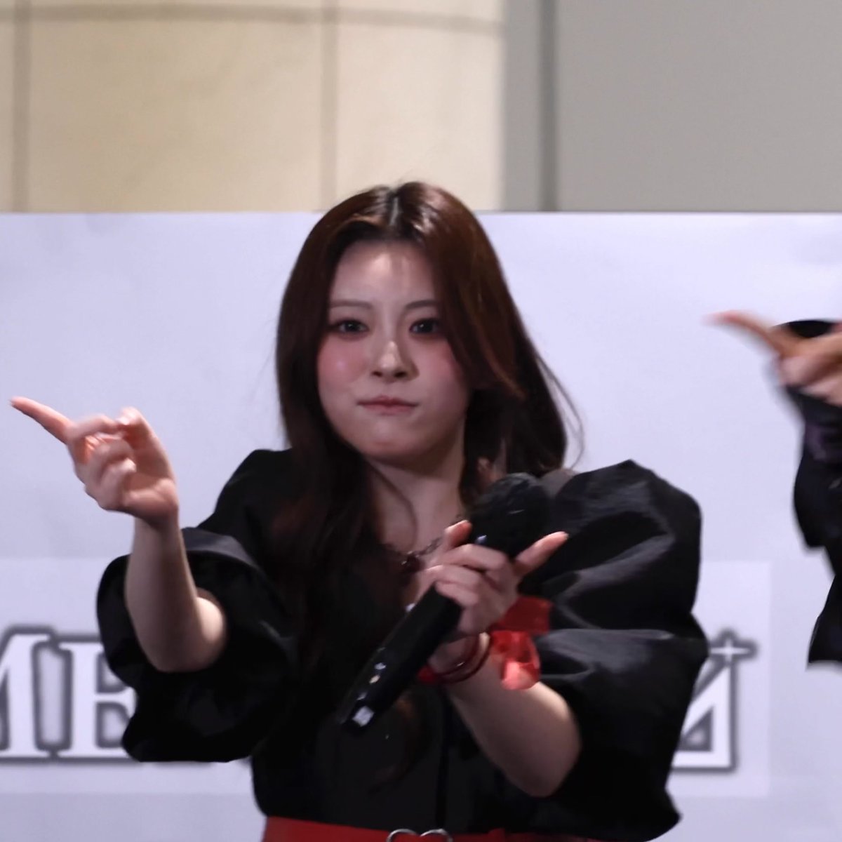 A young Asian woman with long brown hair wearing a black top, holding a microphone and pointing with one hand during what appears to be a fan event or performance.