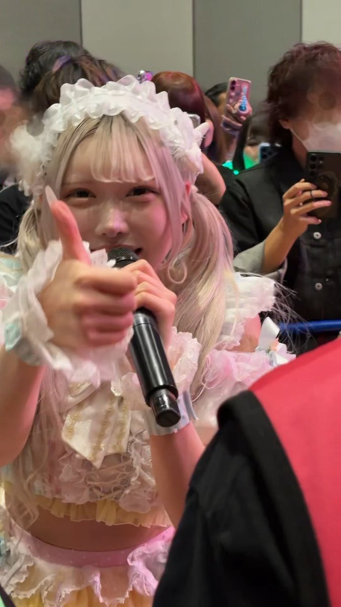 A young woman with blonde and pink hair wearing a frilly white and pink costume and headband, giving a thumbs-up while holding a microphone, surrounded by other people taking photos.