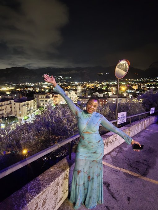 A woman in a flowing light blue and orange patterned dress poses joyfully with arms outstretched at night on a scenic overlook above a city skyline, near a convex traffic mirror.