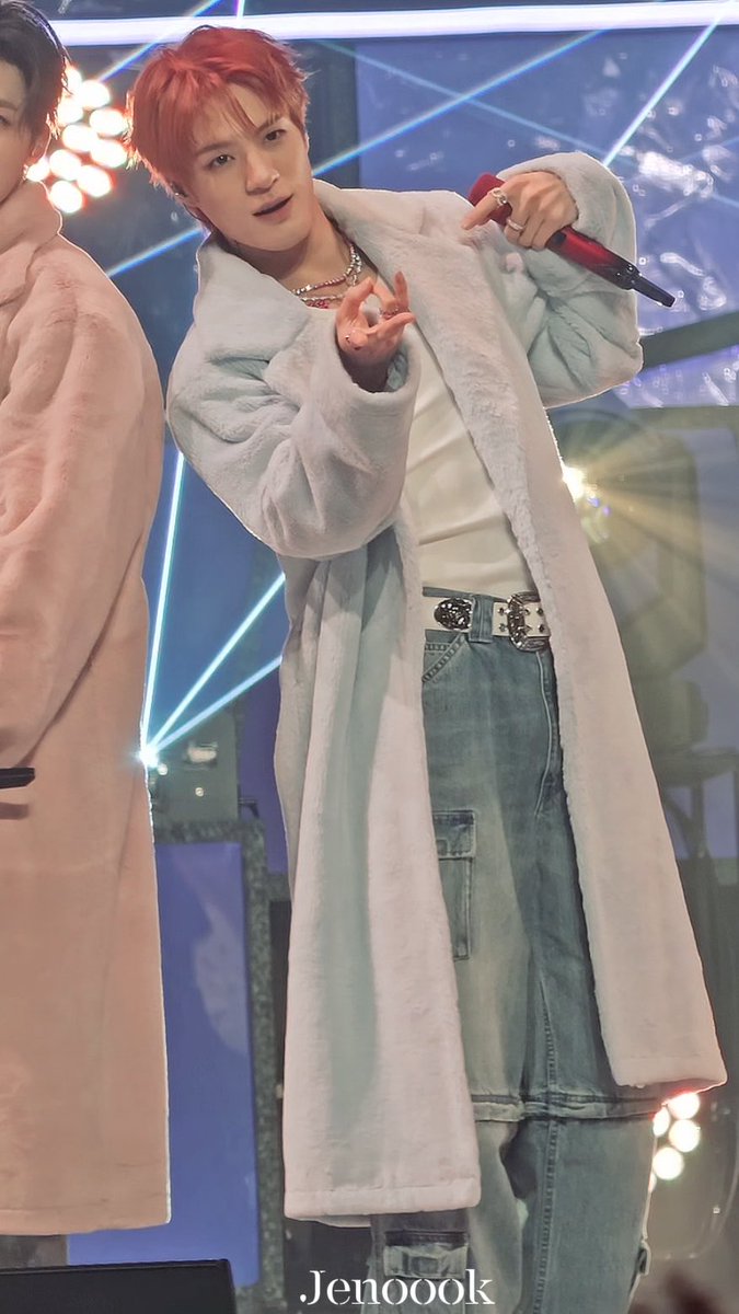 A K-pop performer with reddish-orange hair, wearing a long light blue fuzzy coat and denim cargo pants with a decorated belt, performs on stage holding a red microphone (watermark: Jenooook).