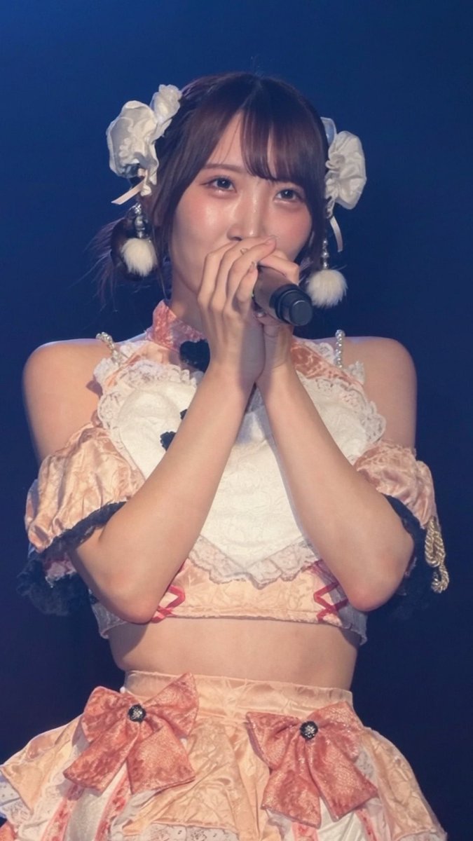 A photo of a Japanese female performer on stage singing into a microphone, wearing an ornate pink and white frilly idol costume with bow hair accessories and a matching skirt.