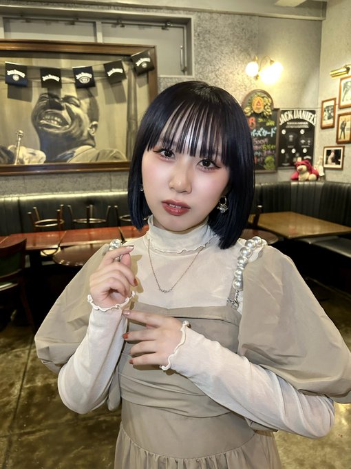 A young woman with short black hair and bangs, wearing a sheer long-sleeved top with pearl details and layered necklaces, poses inside a restaurant decorated with Jack Daniel's decor and a large portrait.