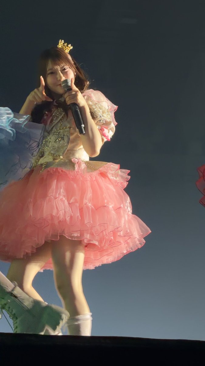 A young woman performs on stage wearing a pink frilly dress and a small gold crown, holding a microphone while making a peace sign and pointing upward.