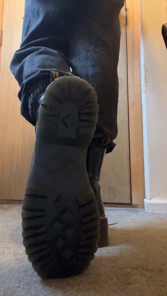A close-up, low-angle shot of the sole of a black work boot showing a deep tread pattern, with the person wearing dark work pants standing on carpet.