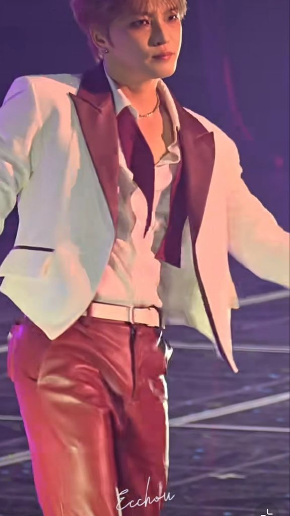 A performer with short hair on stage wearing a white blazer with maroon lapels over a white shirt and maroon leather pants, with the watermark 'Eccho' visible at the bottom.