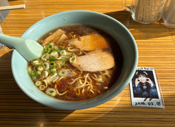 A light blue bowl of ramen with chashu pork, green onions, and bamboo shoots sits on a wooden table next to a small polaroid photo dated '2026.03.08'.