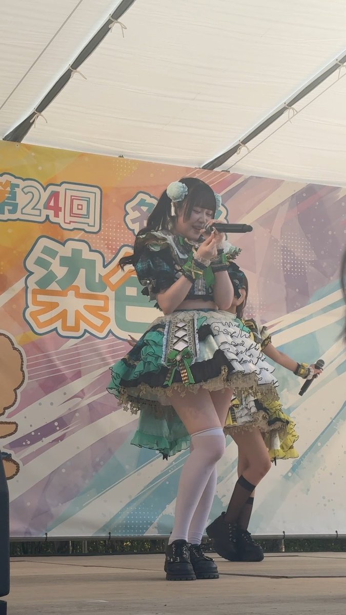 A young performer in an elaborate green and black frilly idol costume sings into a microphone on stage, with a banner behind her displaying Japanese text including '第24回' and '祭'.