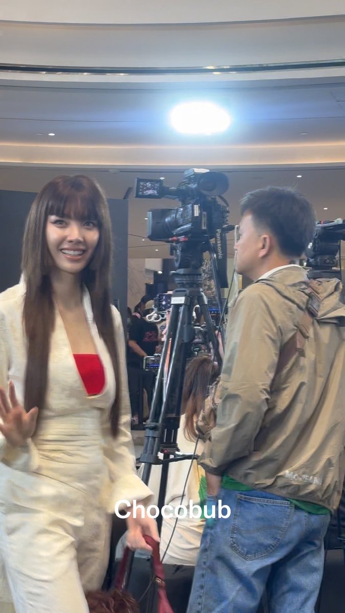 A smiling woman in a white blazer and skirt, wearing a red top underneath, standing near a camera operator and a large broadcast camera; the image is watermarked 'Chocobub'.