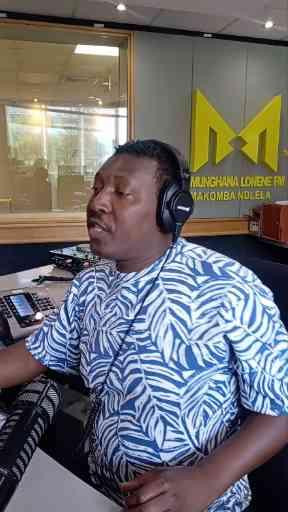 A man wearing headphones and a blue and white leaf-patterned shirt sits at a radio station desk displaying the logo 'MUNGOSHA LOVERE FM YAKOMBA NDOLA'.