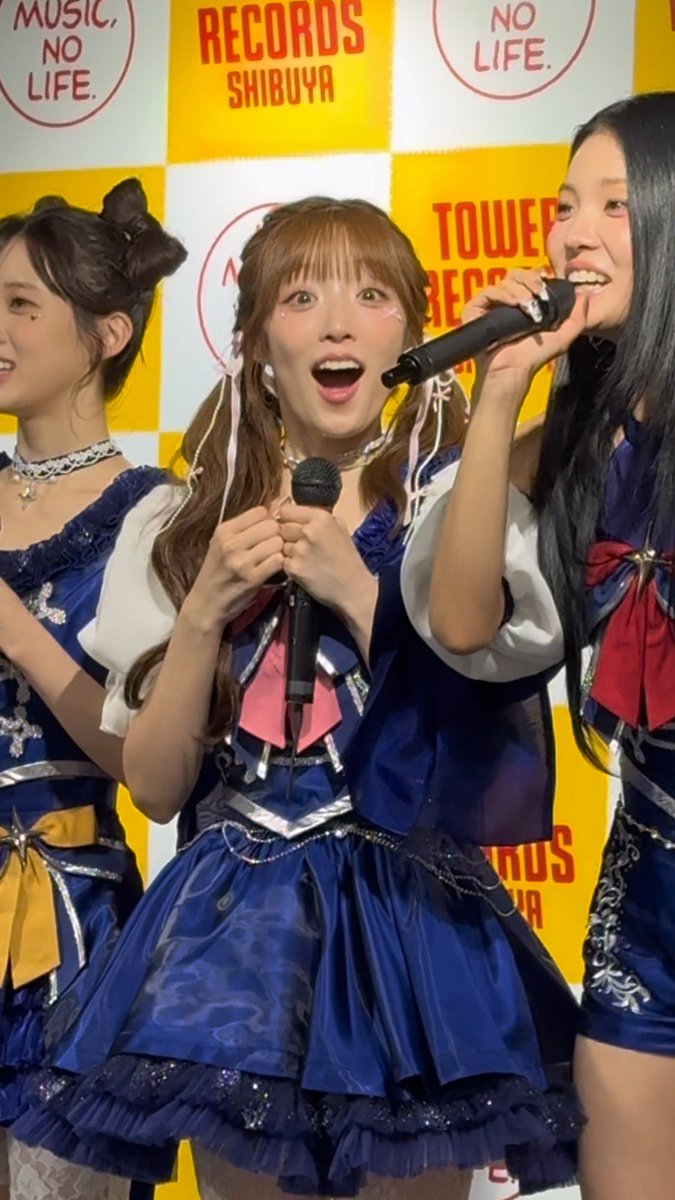 Three young women in matching blue idol-style dresses with pink bows perform at an event, with the center woman singing into a microphone; the background displays 'TOWER RECORDS SHIBUYA' and 'MUSIC, NO LIFE' logos on a yellow and white checkered backdrop.