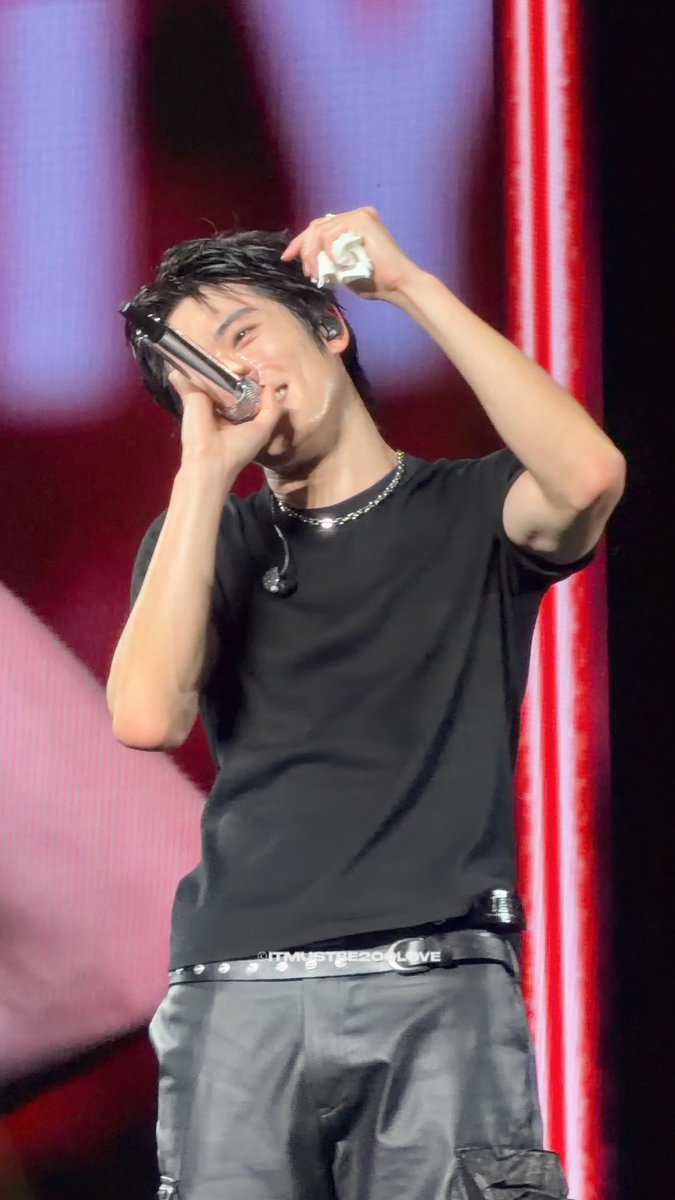 A young male performer on stage singing into a microphone, wearing a black t-shirt with '@ITMUSTBELOVE' text and a belt with '#MUSTBELOVE'.
