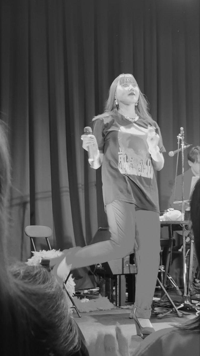 A black and white photo of a female performer on stage, holding a microphone and lifting one leg in a dramatic pose.