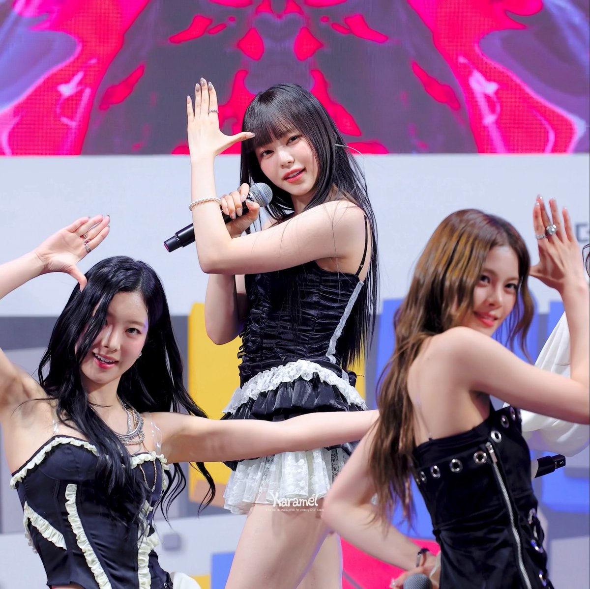 Three female performers are on stage performing, with the central figure singing into a microphone wearing a black ruched top and white skirt; a watermark reading 'Karamel' is visible.