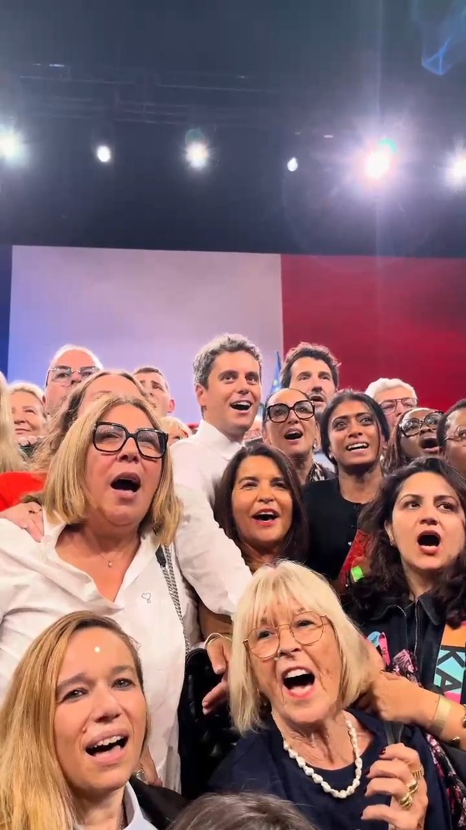A group of people, including a smiling man in a white shirt, pose together with open mouths as if singing or shouting, on a brightly lit stage with a red and white background.
