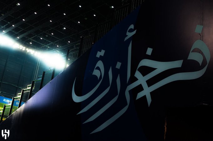 A low-angle shot captures Arabic calligraphy reading "خليجية" (Gulf) on a large blue banner inside a brightly lit stadium. The scene evokes the post's sentiment of life returning to normal, as the stadium setting suggests the resumption of sporting events like the Gulf Cup.