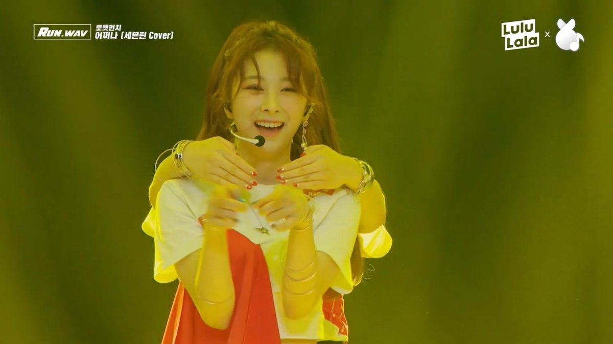 A K-pop performer on stage wearing a headset microphone and white top, forming a heart shape with their hands, with on-screen text 'RUN.WAV' and 'Lovelala (세븐틴 Cover)' and 'LuluLala' branding.