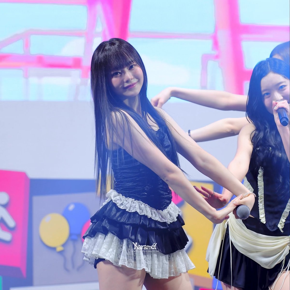 A K-pop performer with long dark hair and bangs wears a black and white ruffled dress, posing on stage with a microphone; a watermark reads 'Karamel Image Hosting for K-pop STAY Girls' Generation'.
