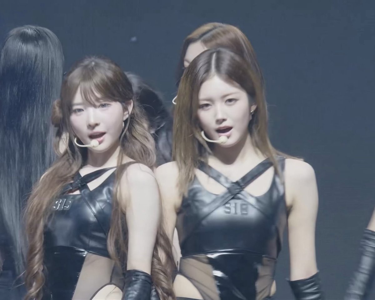 Two female performers on stage in matching black leather-style tops with criss-cross straps and headset microphones.