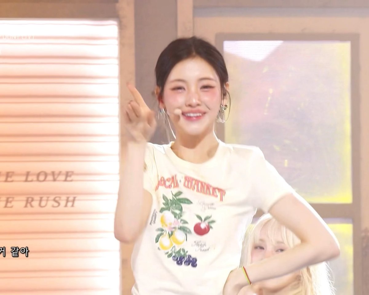 A performer on stage winks and smiles while pointing upwards, wearing a white graphic t-shirt featuring lemons and an apple, with partial text visible on a backdrop behind her.