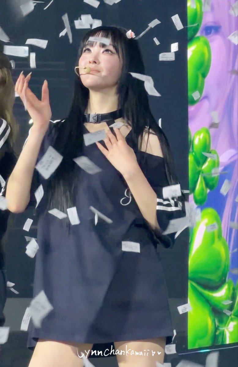 A young woman with long dark hair, wearing a dark off-shoulder top and choker, performs on stage as paper confetti falls around her; watermark reads 'LynnChankawaii'.