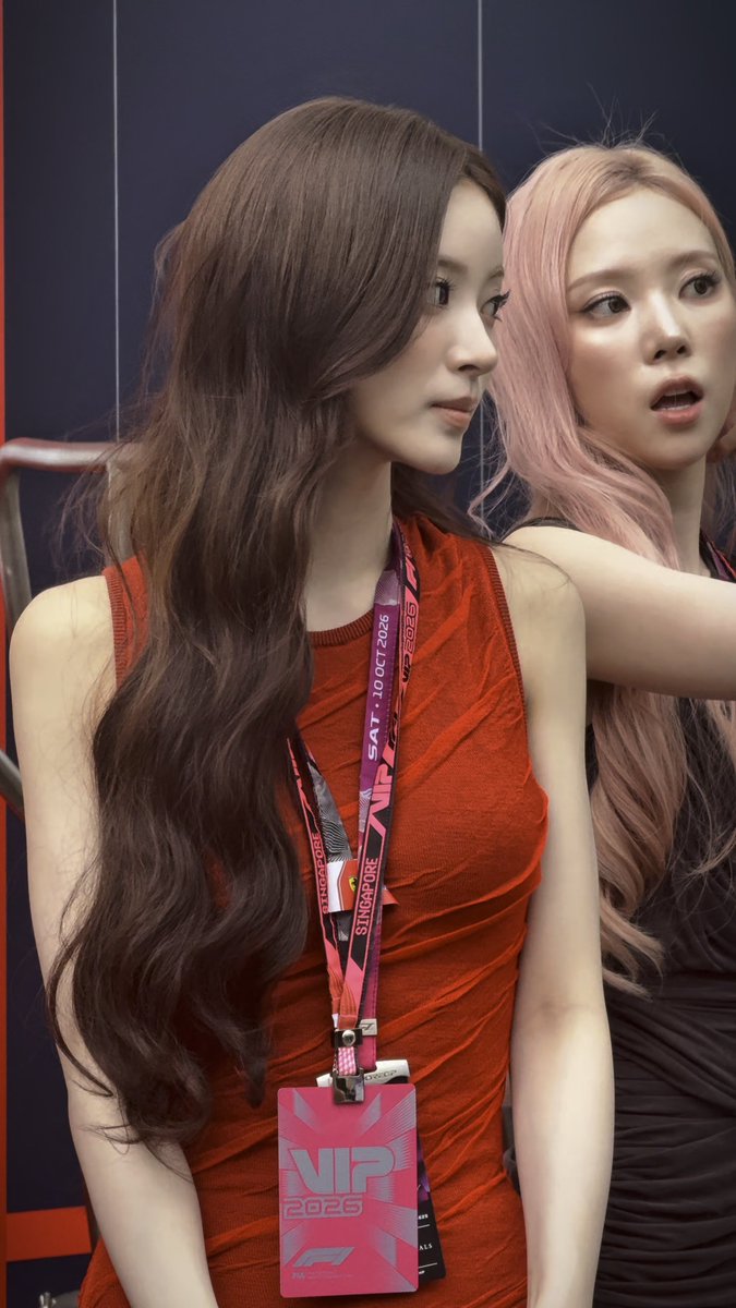 Two young women, one with long dark brown wavy hair wearing a red dress with a VIP pass for the Singapore F1 Grand Prix 2026 (dated SAT, 10 OCT 2026), and another with pink hair, pose together—likely at the event—capturing an excited "Wow" moment.