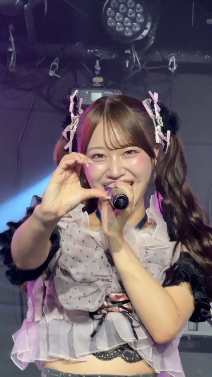 A young woman in a frilly purple and white idol costume makes a heart shape with her hand while singing into a microphone on a stage with theatrical lighting.