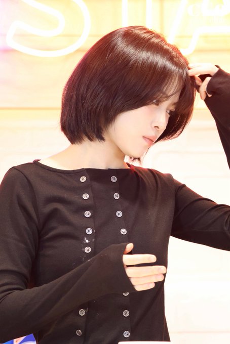 A woman with a sleek short auburn bob haircut wears a black long-sleeved top with double rows of buttons, posing with her hand near her head.