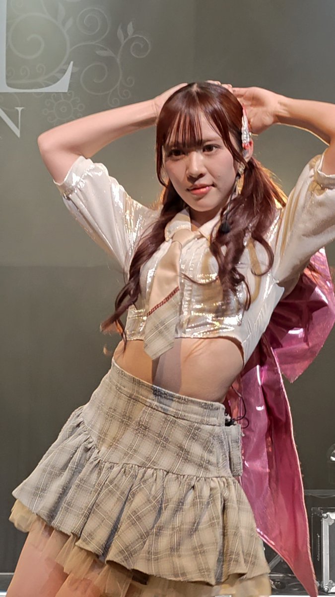 A performer with long brown hair poses on stage, wearing a white and gold cropped shirt with a tie and a plaid skirt, with both hands raised behind her head.