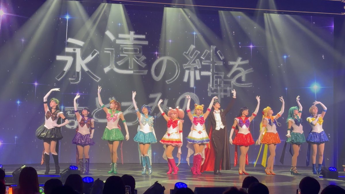 Performers in Sailor Moon costumes and a tuxedo-clad Tuxedo Mask pose on stage with raised arms in front of a backdrop displaying the Japanese text "永遠の輝きを" (eternal brilliance).