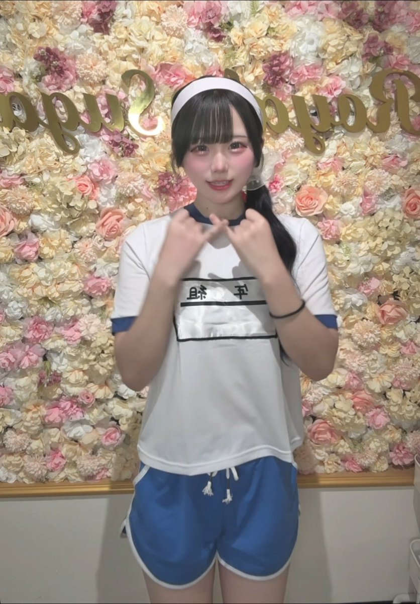 A young woman with dark hair wearing a white headband and a white and blue athletic outfit with Chinese characters makes a heart shape with her hands, standing in front of a wall decorated with pink and white flowers and gold lettering.