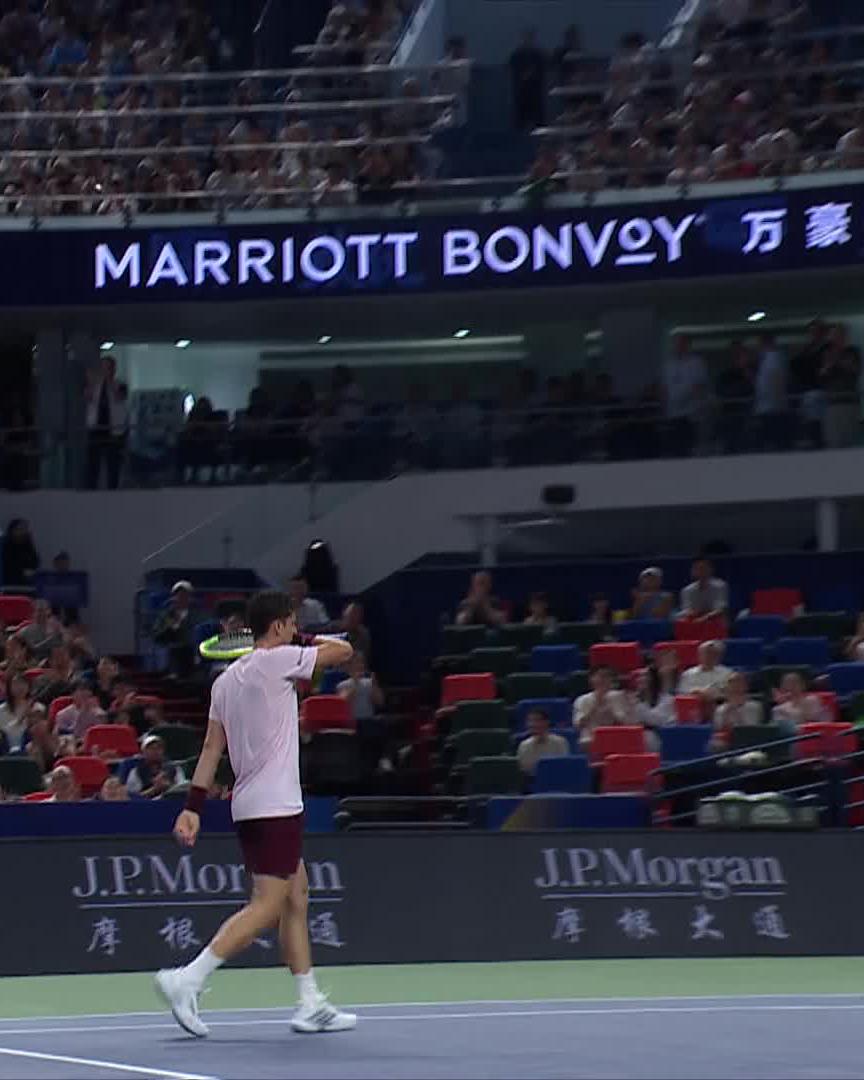 A tennis player in a pink shirt and maroon shorts walks across the court wiping his face with his sleeve, while the crowd looks on from behind a "J.P. Morgan" banner and a "MARRIOTT BONVOY" sign with Chinese characters hangs overhead. The scene captures the post-point moment of the match between Carlos Alcaraz and Juan Manuel Cerúndolo, highlighting the intense atmosphere following the remarkable rally.