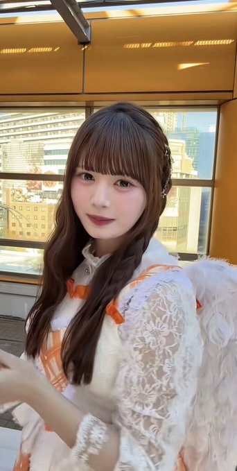 A close-up selfie-style photo of a young woman with long brown hair and bangs, wearing an orange and white lace dress with white feathered wings, with city buildings visible through the window behind her.